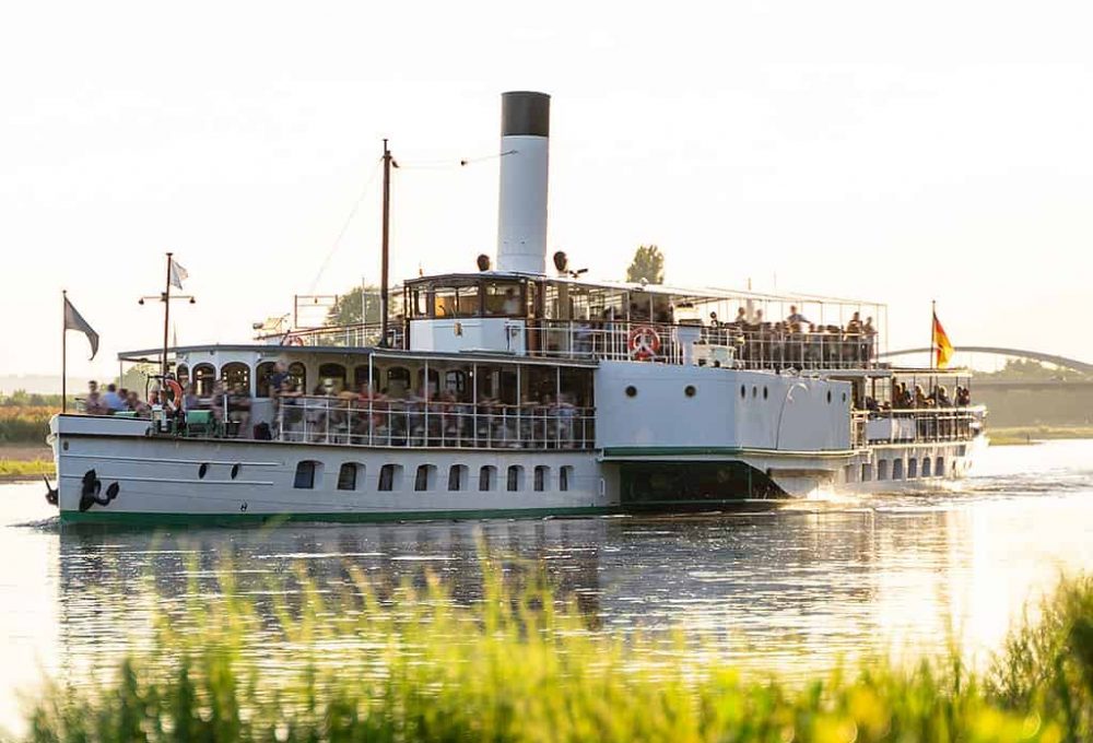 Large retro tourist steamer with a pipe sails along the calm Elbe river Large retro tourist steamer with a pipe is full of people and sails along the calm Elbe river next to a green wild coast on a sunny day at sunset in the city of Dresden. Travel concept